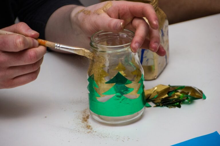 Mão pintando um pote de vidro reaproveitado com tinta verde e dourada
