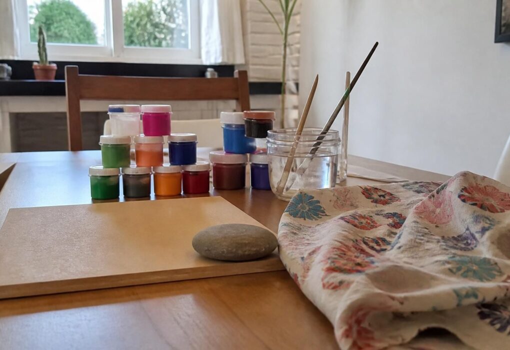 Mesa de artesanato com potes de tinta, pincéis, um pedaço de MDF, um retalho de tecido e uma pedra lisa