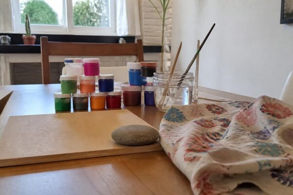 Mesa de artesanato com potes de tinta, pincéis, um pedaço de MDF, um retalho de tecido e uma pedra lisa
